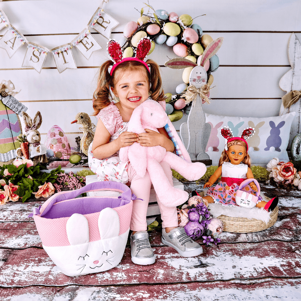 young girl smiling and holding pink plush bunny and sitting in between a doll and easter basket